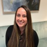 Woman with long, straight brown hair smiling and sitting on a light green sofa with abstract art in the background.