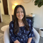 Woman with long dark hair sits in a chair, smiling, wearing a navy blue floral dress in a well-lit room.