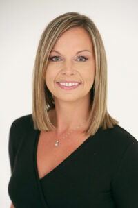 Woman with straight, shoulder-length blonde hair, wearing a black top and a necklace, smiles in front of a plain background.
