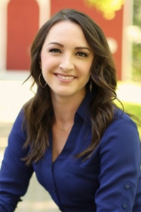 Woman with long brown hair in a blue blouse smiles outdoors with a blurred red and white background.