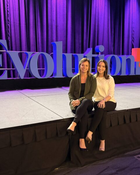 Two women sit on a stage in front of a large "Evolution" sign with purple lighting and a curtain backdrop.
