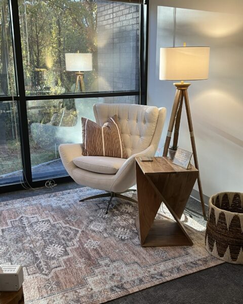 A cream armchair with striped pillows sits by a window, next to a wooden side table and a tripod floor lamp.