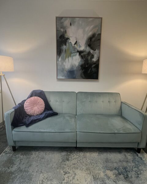 A light blue sofa with a round pillow and blanket sits between two floor lamps below an abstract painting in a modern room.