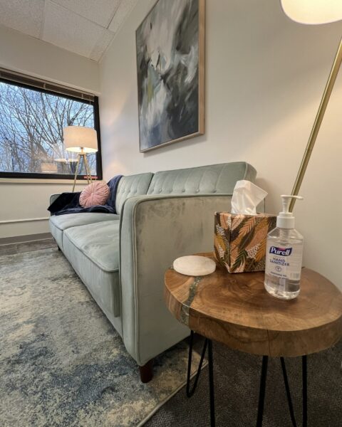 A cozy office with a light green sofa, side table, tissue box, hand sanitizer, lamp, and a window with a tree view.