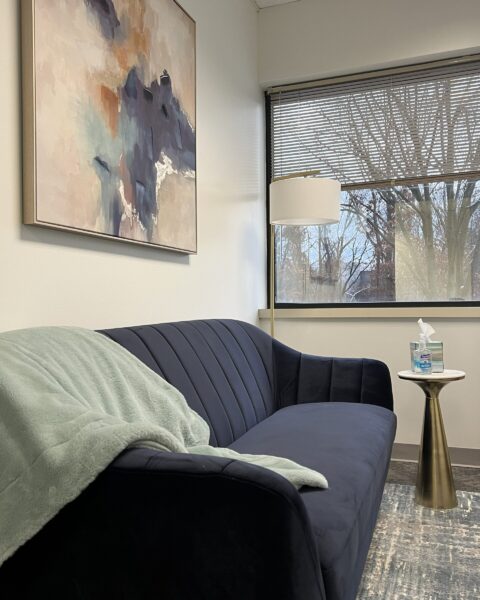A blue couch with a green blanket, side table with tissues, and abstract art in a room with a window.
