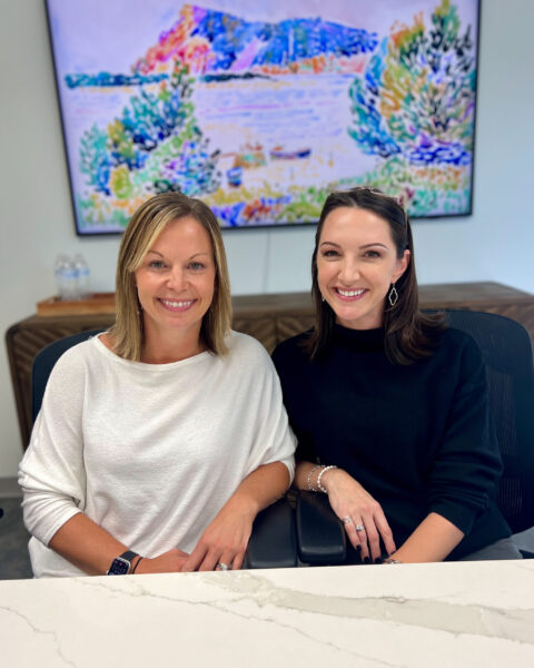 Two women sit at a table, smiling at the camera, with a colorful painting displayed on the wall behind them.