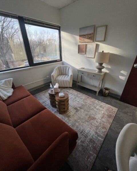 A small office with a red sofa, armchair, side table, lamp, and large window overlooking trees.