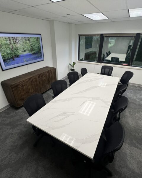 Conference room with a long white table, eight black chairs, a wall-mounted screen, and large windows.