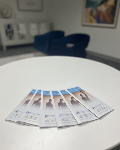 Five brochures are spread out on a round white table in a modern office waiting area with blue chairs.