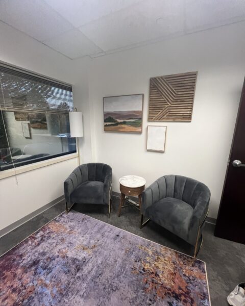 Two gray armchairs and a small round table sit on a colorful rug in a modern office with wall art and a window.