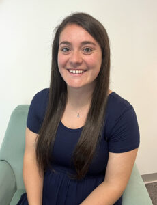 Woman with long brown hair wearing a navy dress sits and smiles at the camera in a light-colored room.