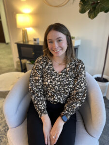 A woman with brown hair sits and smiles, wearing a floral top and black pants in a well-lit office setting.