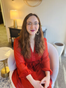 Woman with long brown hair and glasses, wearing a red dress, sitting in a white chair in a well-lit room.