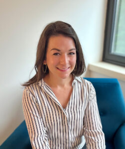 Woman with long brown hair, wearing a white and black striped shirt, sits on a blue chair near a window, smiling at the camera.